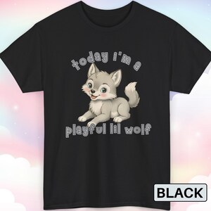 以下が含まれることがあります： 黒いTシャツ。漫画のオオカミのイラストが描かれています。オオカミはグレーと白で、青い目とピンクの鼻をしています。Tシャツのテキストは「today I'm a playful lil wolf」です。