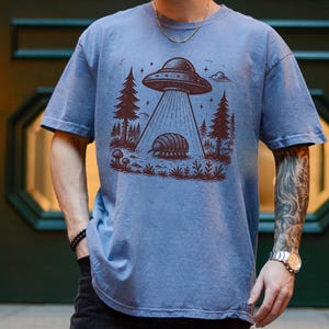 May include: A blue t-shirt with a graphic of a UFO hovering over a pill bug in a forest setting. The graphic is in brown ink.