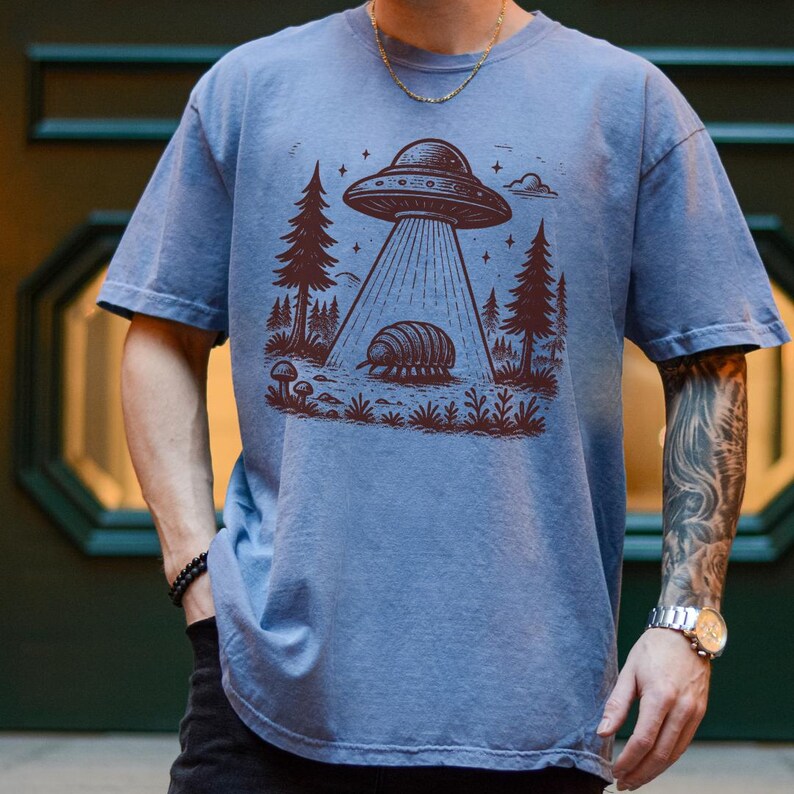May include: A blue t-shirt with a graphic of a UFO hovering over a pill bug in a forest setting. The graphic is in brown ink.