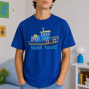 Op de afbeelding: Een koningsblauw T-shirt met korte mouwen voorzien van een grafische illustratie van een blauwe sleepboot met gele accenten, witte reddingsboeien en de tekst "toot toot!" in lichtblauwe letters in het midden van het kledingstuk.