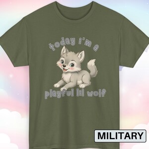 以下が含まれることがあります： オリーブグリーンのTシャツ。漫画のオオカミと「today i'm a playful lil wolf」の文字がプリントされています。オオカミはグレーと白です。Tシャツはクルーネックです。