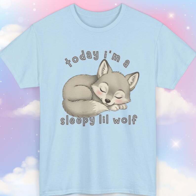 May include: Light blue t-shirt featuring a sleeping gray wolf illustration with the text "today i'm a sleepy lil wolf" in a playful font. The wolf is depicted with closed eyes and a peaceful expression, perfect for casual wear.