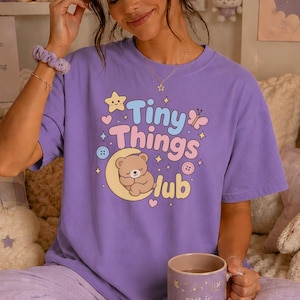 May include: A lavender short-sleeve t-shirt featuring a graphic print with the text Tiny Things Club. The design includes a brown bear sleeping on a yellow crescent moon, surrounded by small stars, hearts, buttons, and a butterfly in pastel colours.