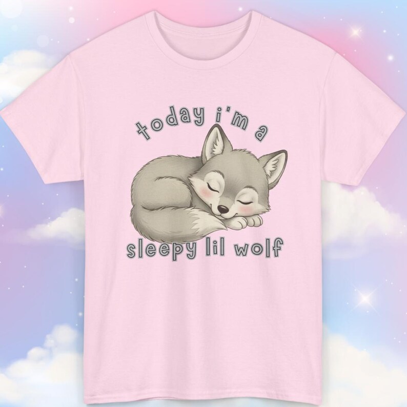 May include: A light pink t-shirt featuring a sleeping gray wolf illustration. The text "today i'm a sleepy lil wolf" is printed above and below the wolf. The shirt has short sleeves and a crew neck.