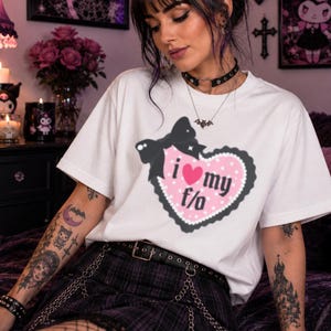 May include: A white short-sleeved t-shirt featuring a graphic of a pink heart with white polka dots, a black bow, and a black lace-style border. The text "i heart my f/o" is printed in the center of the heart with a pink heart icon for the word heart.