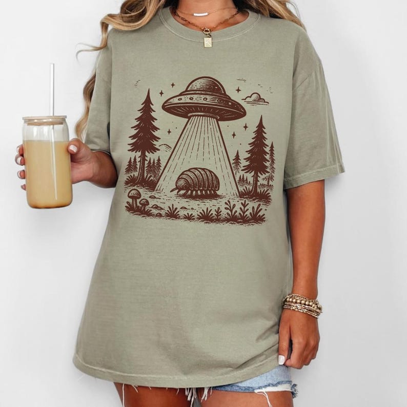 May include: A light green t-shirt with a brown graphic of a UFO hovering over a pillbug in a forest setting.