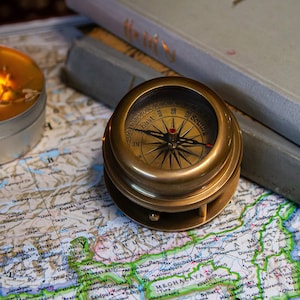 Może przedstawiać: Kompas mosiężny z czarną igłą i białą tarczą z czarnymi oznaczeniami leży na mapie z zapaloną świecą w tle.