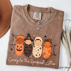 May include: Espresso-colored t-shirt featuring a Halloween-themed graphic of four babies in costumes, including a pumpkin, witch, ghost, and candy corn. The text "Caring for the Spookiest Littles" is printed below the graphic. A silver necklace is draped across the shirt.