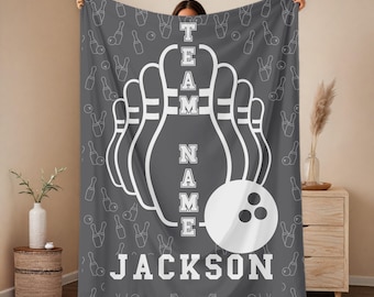 May include: A charcoal grey fleece blanket featuring a repeating white outline pattern of bowling pins and balls. The center displays a large white bowling pin graphic with the vertical text TEAM NAME and the name JACKSON printed below in white.