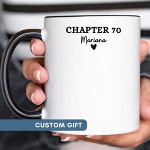 May include: A white ceramic mug with a black handle and rim. The front of the mug features the black text "CHAPTER 70" followed by the name "Mariana" in a script font and a small black heart icon centered below the name.
