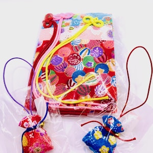 May include: A stack of colourful Japanese fabric with a red background and circular patterns. Several small, decorative pouches in red and blue, each with a bell and a loop, are visible. The pouches are packaged in clear plastic.