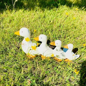 May include: A family of four white ceramic ducks with yellow beaks and feet, arranged in a row on green grass. The ducks decrease in size, from front to back. Yellow wildflowers are scattered around the ducks, adding a touch of colour.