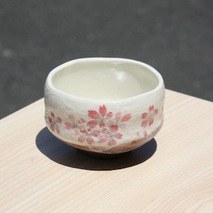 May include: A ceramic bowl with a cream-colored interior and a textured, white exterior. The bowl is adorned with delicate pink cherry blossom designs around the rim. The base has a brown, earthy tone, and the bowl sits on a light-colored wooden surface.