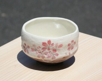 May include: A ceramic bowl with a cream-colored interior and a textured, white exterior. The bowl is adorned with delicate pink cherry blossom designs around the rim. The base has a brown, earthy tone, and the bowl sits on a light-colored wooden surface.