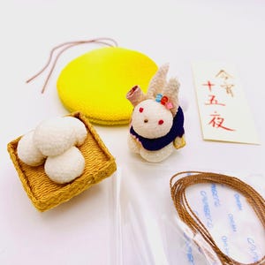May include: A selection of Japanese craft items. Features a yellow circular cushion, a rabbit doll with a blue collar, a woven basket with three white mochi, and a card with Japanese text. Also includes brown string and a clear plastic bag.