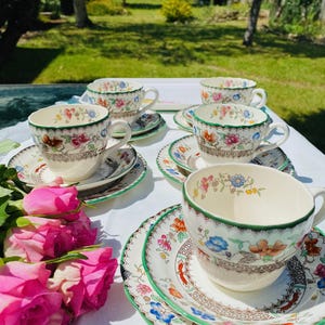 May include: A set of vintage teacups and saucers with floral patterns. The cups and saucers have a white base with colorful flowers and a green trim. A bouquet of pink roses is in the foreground. The scene is set outdoors on a white tablecloth.