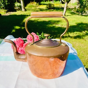May include: A vintage copper and brass kettle with a wooden handle. The kettle has a rounded body and a spout. The handle is arched. The kettle is on a white tablecloth with pink roses in front of it.