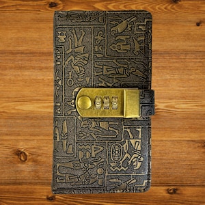 Könnte beinhalten: Ein schwarzes Lederjournal mit einem goldenen Zahlenschloss und einem geprägten Muster aus alten ägyptischen Hieroglyphen.