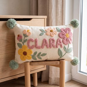 May include: A rectangular off-white throw cushion featuring the name CLARA embroidered in raised pink yarn. The cushion is decorated with punch needle floral motifs in yellow, pink, and peach, accented by sage green leaves and four sage green pom-poms.