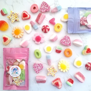May include: A collection of colorful candies in various shapes, including hearts, stars, flowers, and rings, scattered on a white marble surface. Two pink bags filled with assorted sweets are also present. Some candies are coated in sprinkles, and others have a jelly-like texture.