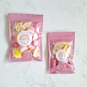 May include: Two pink cellophane bags filled with assorted candies. The bags contain star-shaped, heart-shaped, and flower-shaped sweets in pink, yellow, and white. Each bag has a circular sticker with a rainbow and the text "Olivia's 7th Birthday."