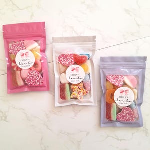 May include: Three clear plastic bags filled with colorful candies. Each bag has a label that reads "Emily's hen do 25.02.2026". The bags are pink, clear, and lavender, and are arranged on a white marble surface.