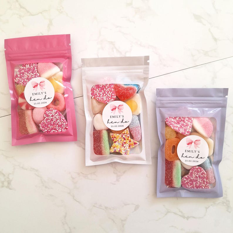 May include: Three clear plastic bags filled with colorful candies. Each bag has a label that reads "Emily's hen do 25.02.2026". The bags are pink, clear, and lavender, and are arranged on a white marble surface.