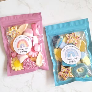 May include: Two clear plastic bags filled with colorful candies. One bag is pink and the other is blue. Each bag has a circular sticker with a rainbow and the text "Thank you for celebrating" and a birthday message.