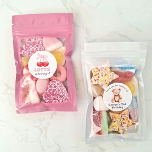 May include: Two clear cellophane bags filled with colorful candies. One pink bag has a sticker that says "Lottie is turning 8" with cherry graphics. The other bag is clear with a sticker that says "Thank you for celebrating" and "George's 2nd Birthday".