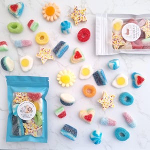 May include: An assortment of colorful gummy candies in various shapes, including hearts, stars, flowers, and rings. Some candies are coated in sugar. Two bags of candies are visible, one with a label that reads "Freddie's 1st Birthday".