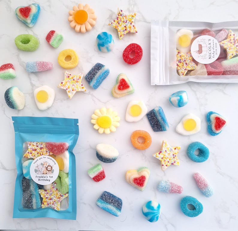 May include: An assortment of colorful gummy candies in various shapes, including hearts, stars, flowers, and rings. Some candies are coated in sugar. Two bags of candies are visible, one with a label that reads "Freddie's 1st Birthday".