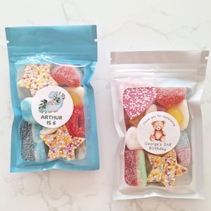 May include: Two clear cellophane bags filled with colorful candies. One bag is light blue and has a sticker that says "ARTHUR IS 6". The other bag is clear and has a sticker that says "George's 2nd Birthday".