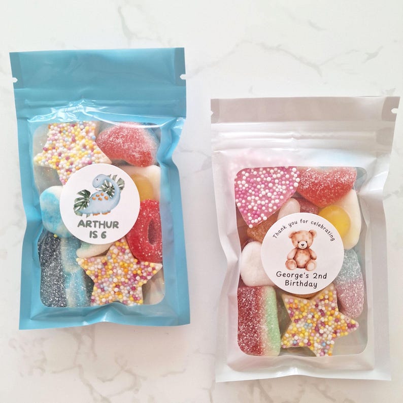 May include: Two clear cellophane bags filled with colorful candies. One bag is light blue and has a sticker that says "ARTHUR IS 6". The other bag is clear and has a sticker that says "George's 2nd Birthday".