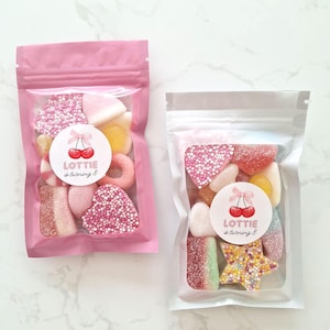 May include: Two clear cellophane bags filled with colorful candies. One bag is pink, the other is clear. Each bag has a round sticker with cherries and the text "Lottie is turning 8". The candies include hearts, rings, and star shapes.