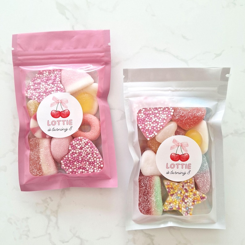 May include: Two clear cellophane bags filled with colorful candies. One bag is pink, the other is clear. Each bag has a round sticker with cherries and the text "Lottie is turning 8". The candies include hearts, rings, and star shapes.
