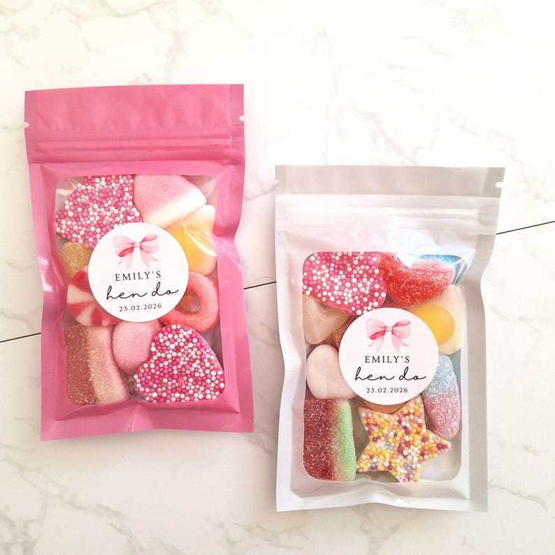 May include: Two clear plastic bags filled with assorted candies. One bag is pink, the other is clear. Each bag has a circular sticker that reads "Emily's hen do 23.02.2026" with a bow graphic. The candies are various shapes and colors.