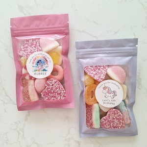 May include: Two clear plastic bags filled with colorful candies. One bag is pink and the other is lavender. Each bag has a circular sticker with text and a graphic. The pink bag says "Aubree" and the lavender bag says "Leni's 4th Birthday".