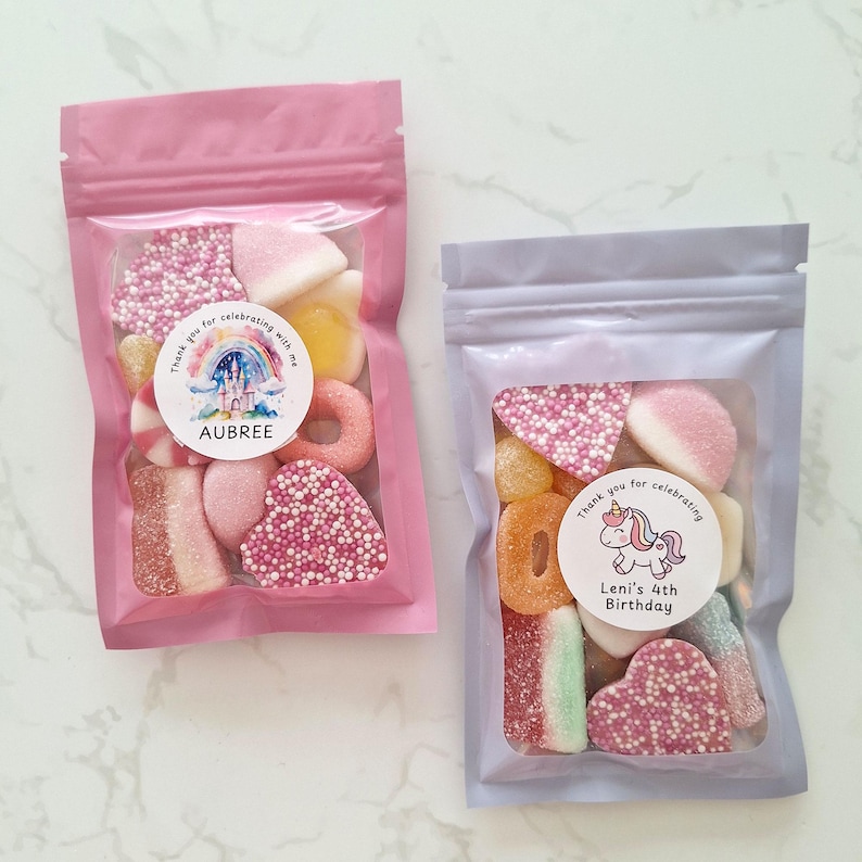 May include: Two clear plastic bags filled with colorful candies. One bag is pink and the other is lavender. Each bag has a circular sticker with text and a graphic. The pink bag says "Aubree" and the lavender bag says "Leni's 4th Birthday".