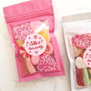 May include: Two pink cellophane bags filled with assorted candies. One bag features a round sticker that reads "Elle's hen party" in red text. The candies include heart-shaped sweets covered in pink sprinkles, and other colorful gummy treats.