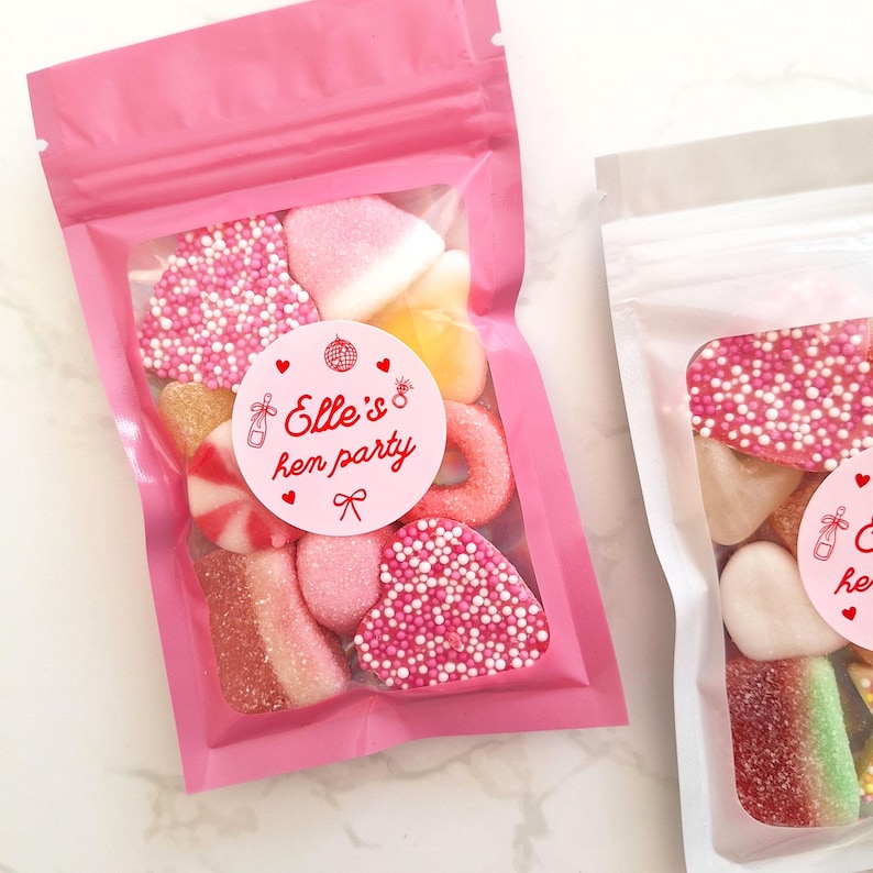 May include: Two pink cellophane bags filled with assorted candies. One bag features a round sticker that reads "Elle's hen party" in red text. The candies include heart-shaped sweets covered in pink sprinkles, and other colorful gummy treats.
