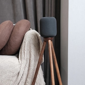 Può includere: Un altoparlante cilindrico grigio scuro su un supporto a treppiede in legno. L'altoparlante è appoggiato su una coperta bianca con cuscini marroni sullo sfondo.