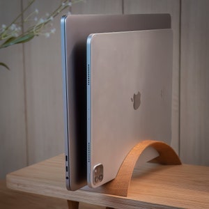 May include: A silver laptop and tablet are stacked on a wooden stand. The laptop is closed and the tablet is open, showing the Apple logo. The stand is made of light brown wood and has a curved shape.