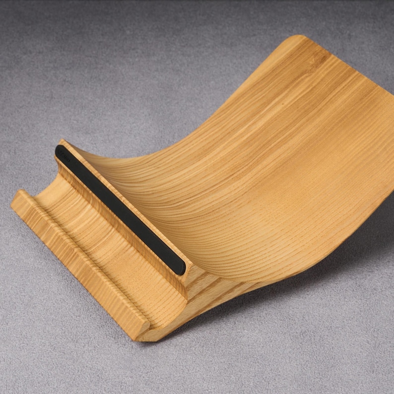 Op de afbeelding: Een houten laptopstandaard met een gebogen ontwerp en een zwarte rubberen strip langs de bovenrand. De standaard is gemaakt van licht hout met een natuurlijke afwerking.