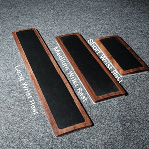 Op de afbeelding: Drie houten polsleuningen met zwarte stoffen bovenkanten. De polsleuningen zijn gelabeld met "Long Wrist Rest", "Medium Wrist Rest" en "Short Wrist Rest".