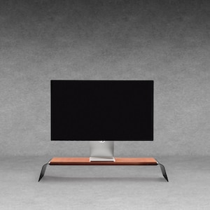 Könnte beinhalten: Ein Computermonitor mit schwarzem Bildschirm steht auf einem Holzständer mit gebogenen schwarzen Metallbeinen. Der Monitor hat einen silbernen Sockel. Der Ständer steht vor einem grauen Hintergrund.