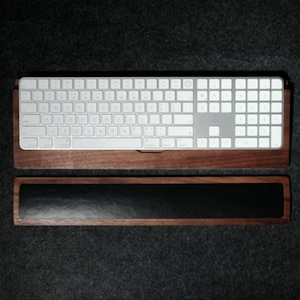 Puede incluir: Una bandeja de teclado de madera con un reposamuñecas de cuero negro. La bandeja está diseñada para sostener un teclado inalámbrico y elevarlo a una altura de escritura cómoda.