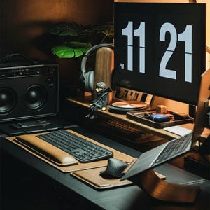 May include: A dark brown wooden desk with a black laptop, a brown leather keyboard wrist rest, a black keyboard, a black speaker, a black and white clock displaying 11:21 PM, a brown wooden stand, and a small figurine.