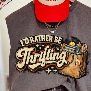 May include: A gray sleeveless t-shirt with the text "I'd Rather Be Thrifting" in a retro font. The graphic includes a shopping bag with a record and other items. A white and red baseball cap with "Darlin'" is above the shirt.