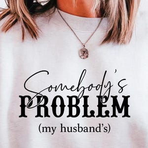 Może przedstawiać: Jasnoszara bluza z napisem "Somebody's PROBLEM (my husband's)" w kolorze czarnym. Bluza ma swobodny styl, odpowiednia do noszenia na co dzień.