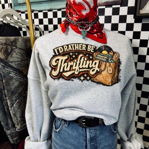 Peut inclure: Un sweat-shirt gris clair avec le texte "I'd Rather Be Thrifting" dans une police r&eacute;tro, avec un graphisme d'un sac de shopping rempli de disques. Le sweat-shirt est associ&eacute; &agrave; un short en jean et une ceinture noire.